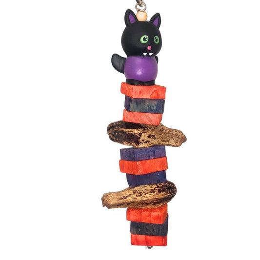 A Halloween-themed wooden bird toy featuring a black cat figurine with green eyes and a purple belly perched atop stacked wooden blocks in orange, purple, and black, with two natural mahogany pads

